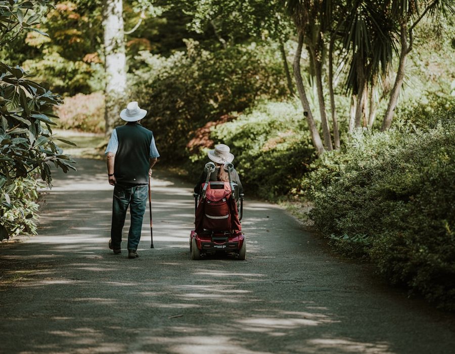 Two people out together on a shaded park path, one walking with a cane and one using a mobility scooter